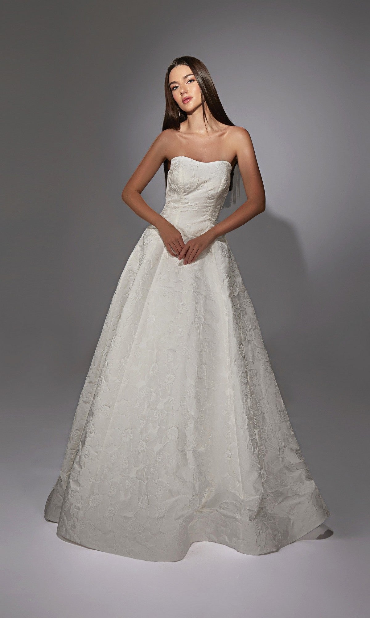 In a studio, a woman wears the Alyce 7155 long white dress in ivory, featuring a strapless design and delicate floral jacquard fabric that creates an elegant, floor-length silhouette.